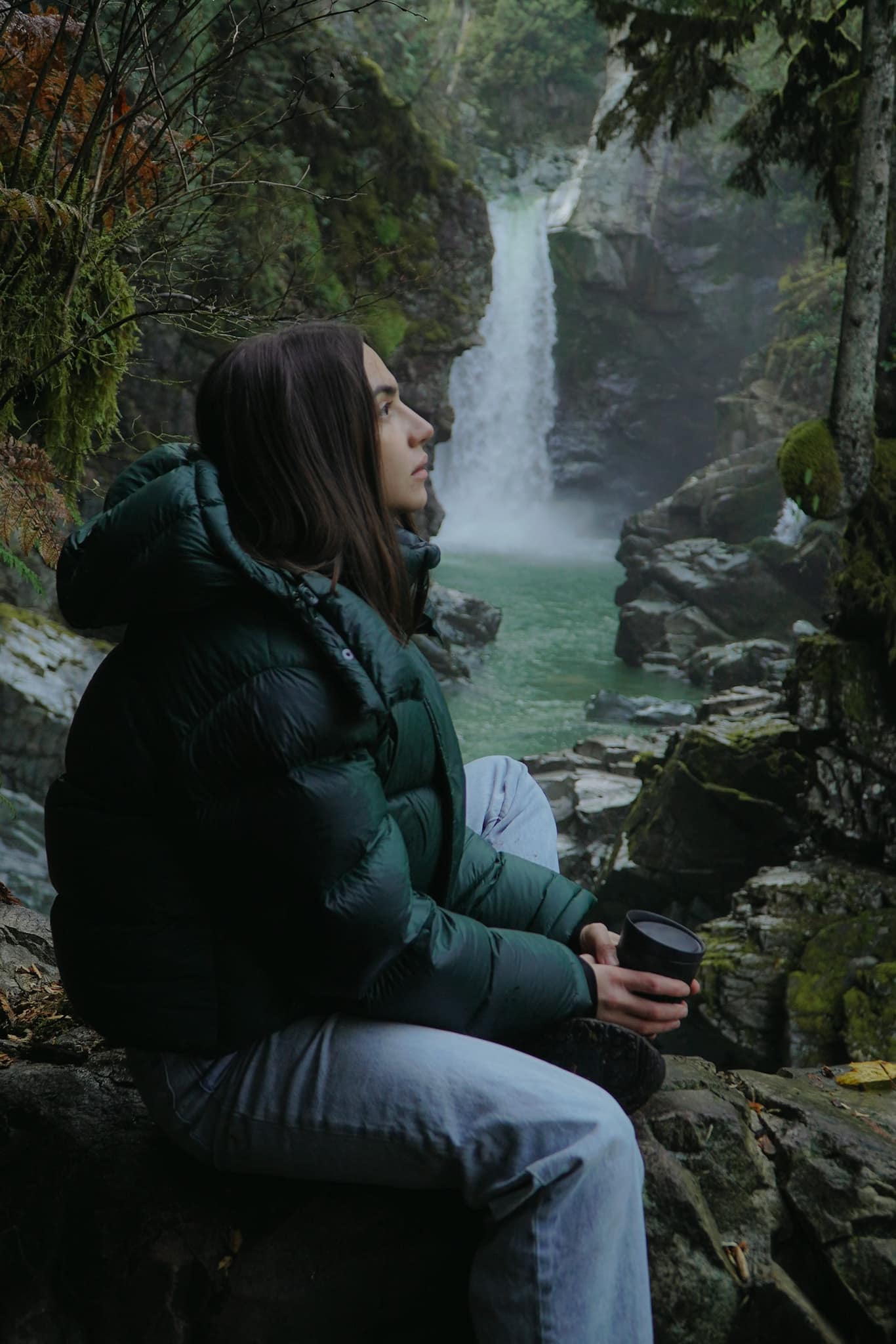 Mariia sitting on the rocks below a rainforest waterfall, a cup in her hands