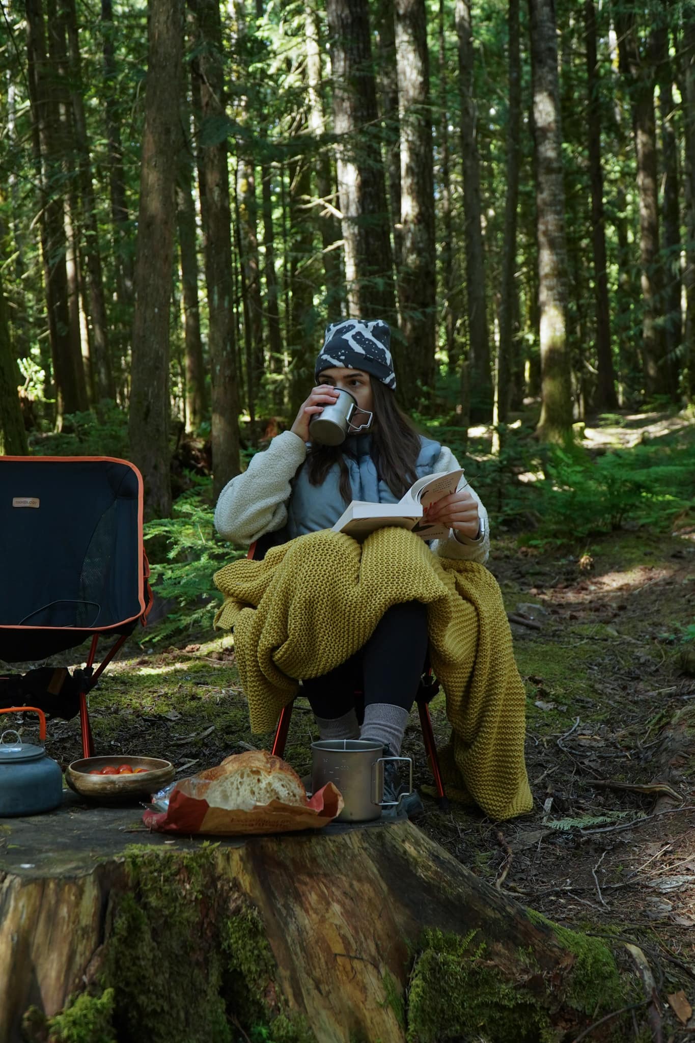 Reading under a knitted blanket at a forest camp, coffee in hand, bread on a stump