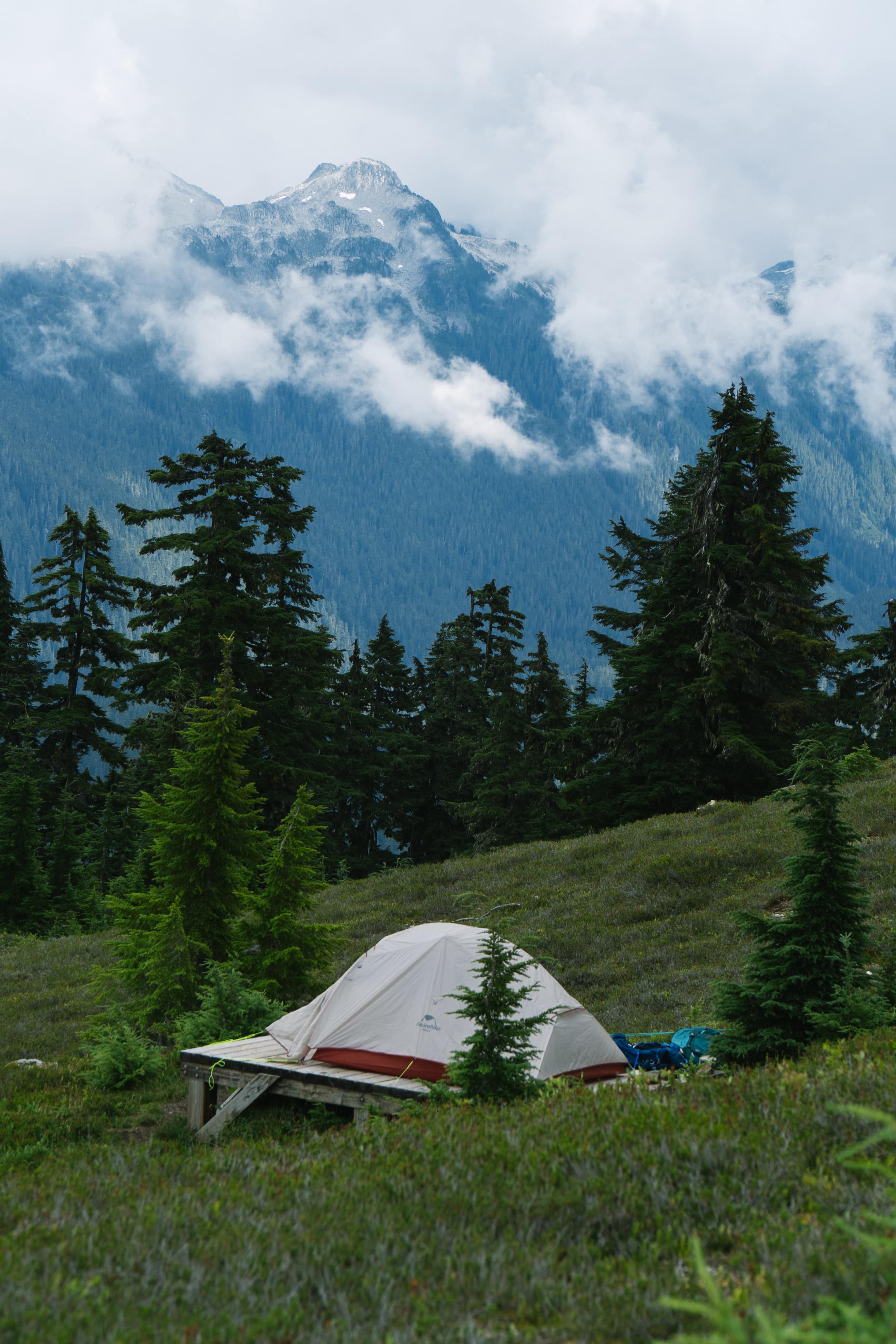 A Naturehike tent on a wooden platform in an alpine meadow, clouds on the peaks