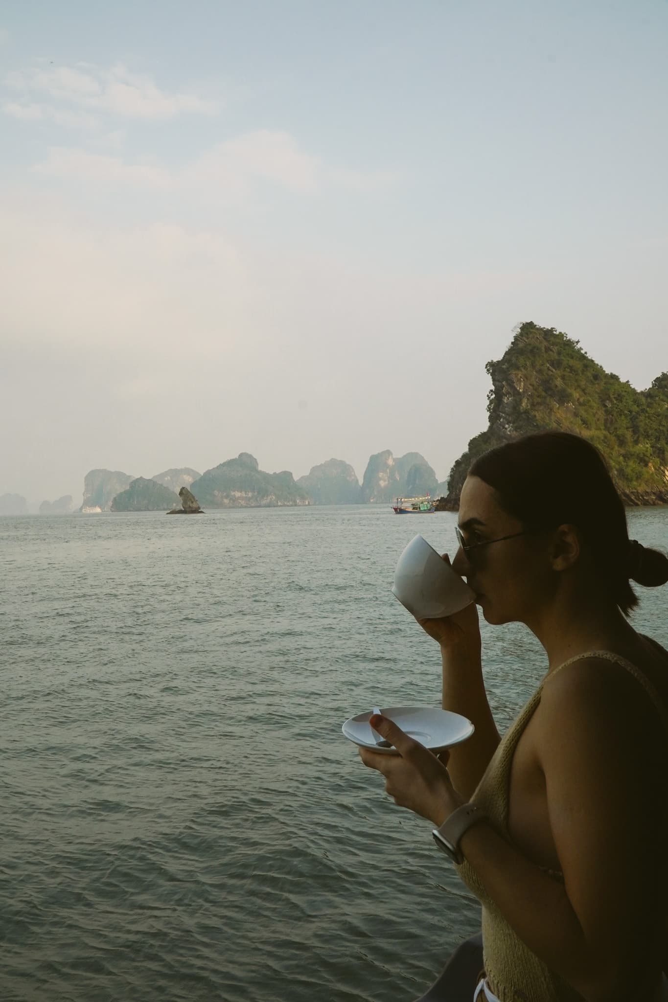 Mariia drinking coffee on deck, limestone islands fading into the haze behind